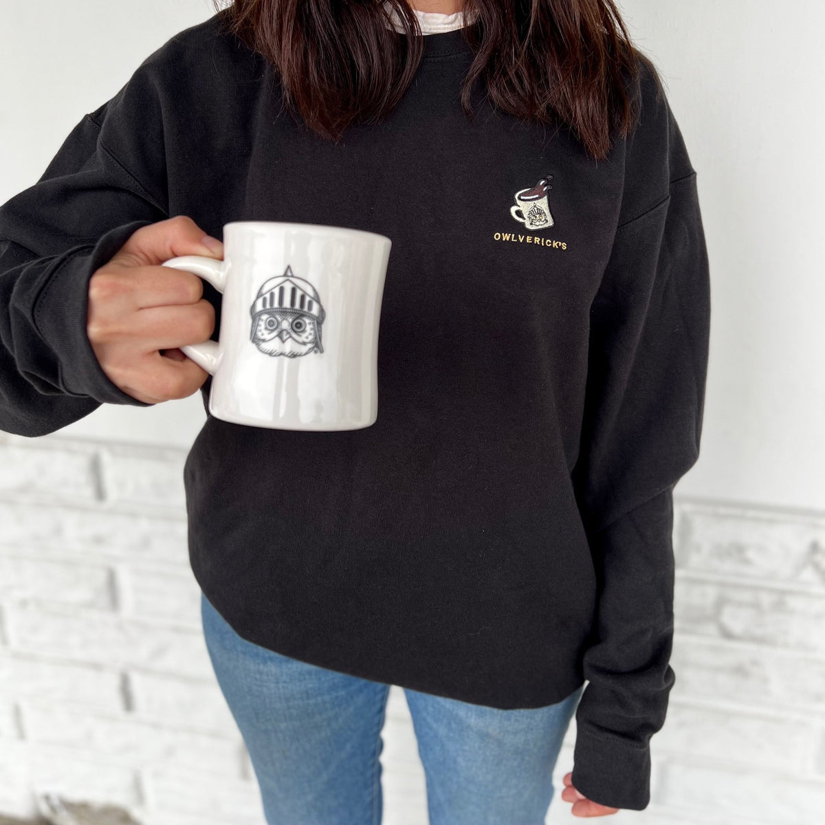 A person wearing a black crewneck sweatshirt with a small gold and white owl graphic on the left chest, holding a white ceramic mug with a black owl illustration. The background is a white brick wall.
