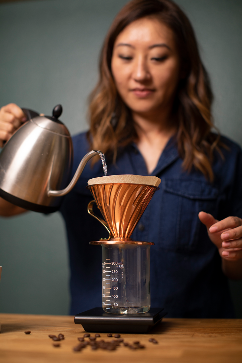 A woman in a navy blue shirt pours water into a copper cone filter, with coffee beans scattered on a wooden table.