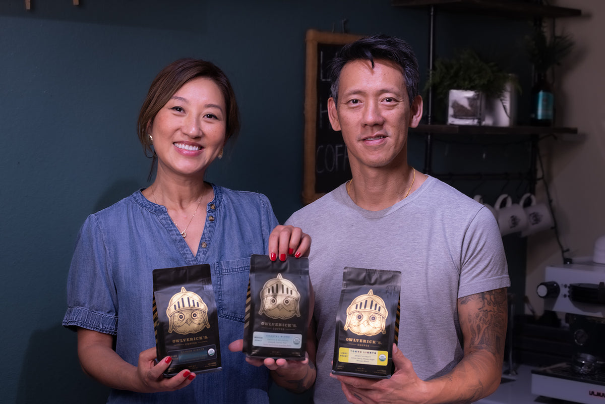 A smiling woman in a blue top and a man in a gray t-shirt stand together holding three bags of coffee from the brand Owlvericks. They are posed in front of a dark teal wall with shelves in the background.