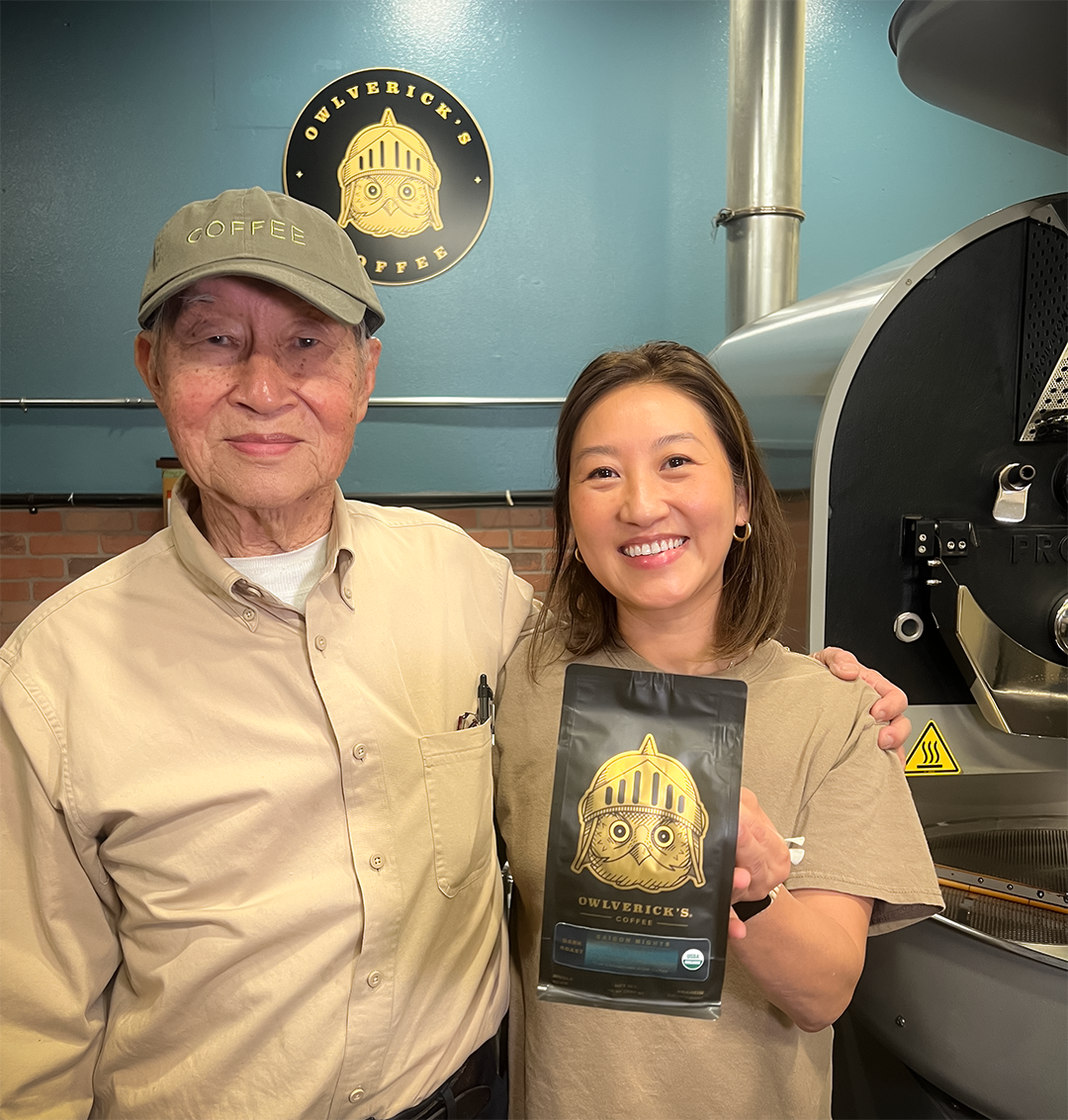 An older man and a woman stand in a coffee roasting room, smiling and holding a black bag of Owlverick's  Whole Bean coffee with a gold knight logo. The background features a teal wall with a circular owl logo and a large coffee roaster.