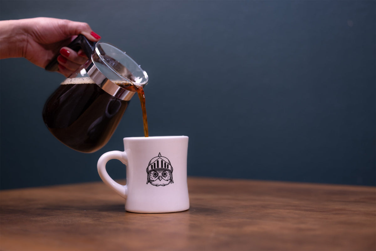 A hand with red nail polish pours dark coffee from a glass carafe into a white ceramic mug featuring a black graphic of an owl in a knight helmet.