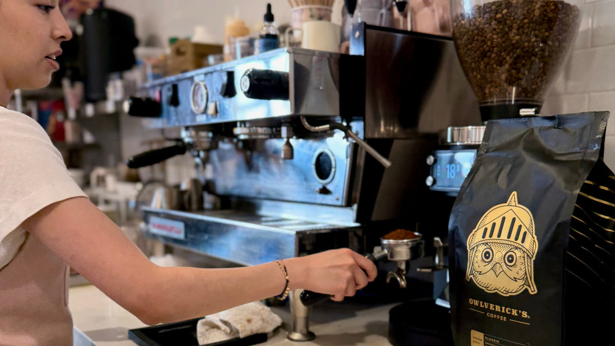 A barista prepares coffee at a counter with a stainless steel espresso machine, coffee grinder, and a black bag of Owlverick's coffee beans featuring a gold owl logo in the foreground.