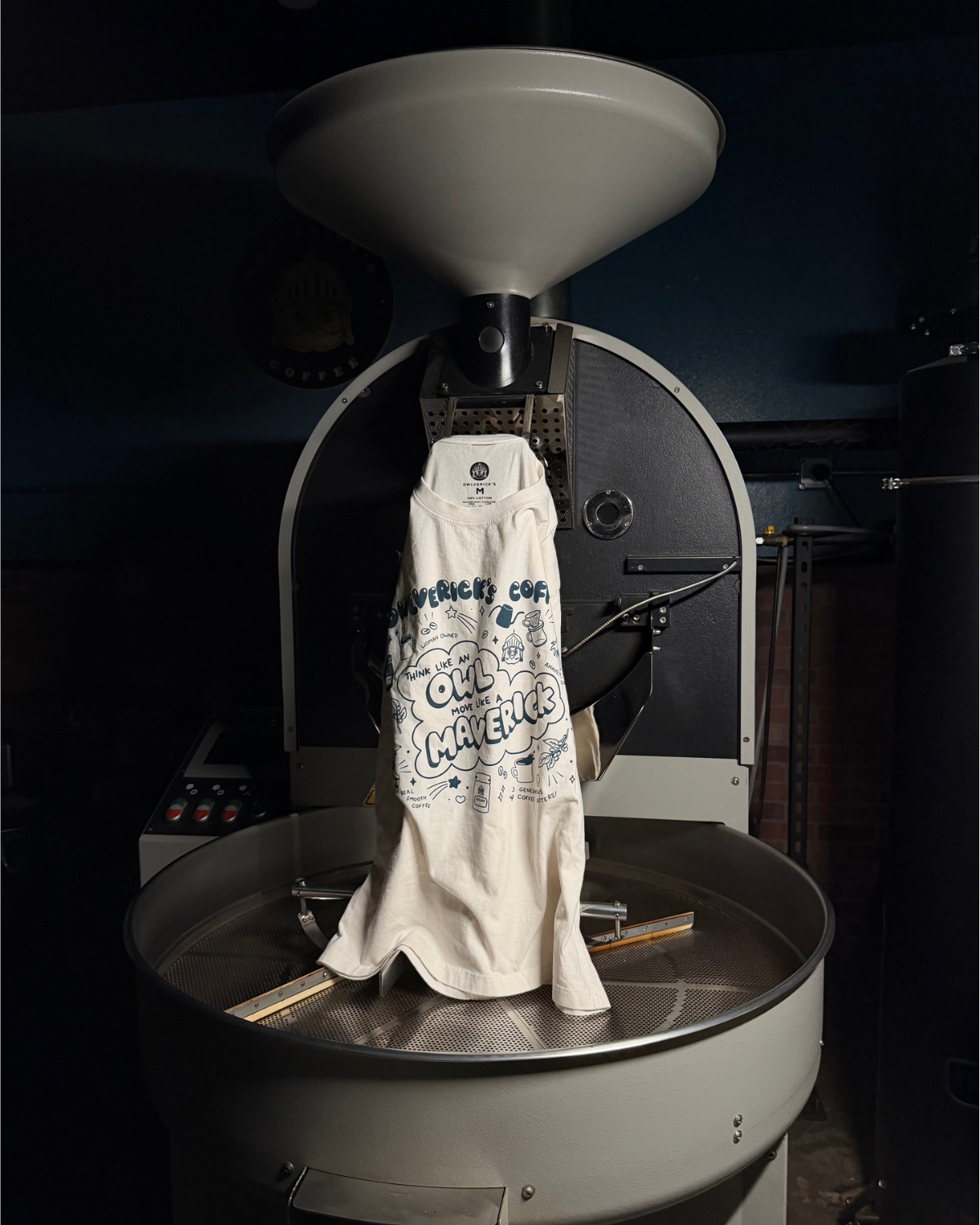 A Vintage White cotton t-shirt with blue graphic text reading 'Owlverick's Coffee' and 'Think Like an Owl, Move Like a Maverick" is draped over a large metal coffee roaster inside a dark, industrial setting.