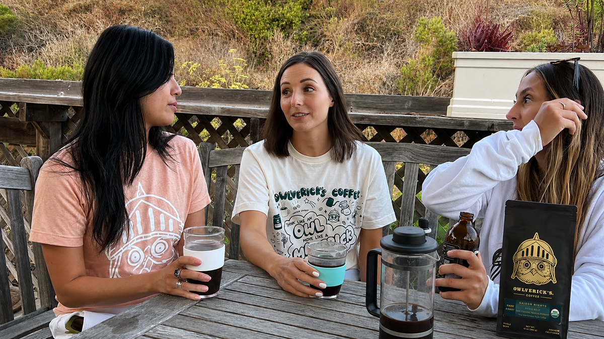 Three women sitting outdoors at a wooden table having coffee, with a French press, mugs, and a bag of coffee beans in front of them. 