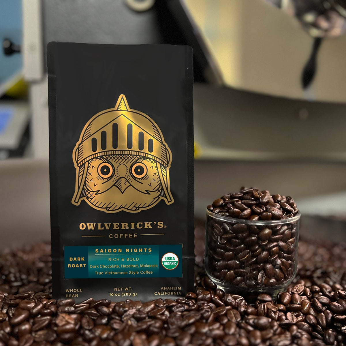 A bag of Owlverick's Coffee 'Saigon Nights' dark roast coffee stands upright on a bed of loose coffee beans, with a small glass jar of roasted beans and a coffee machine blurred in the background.