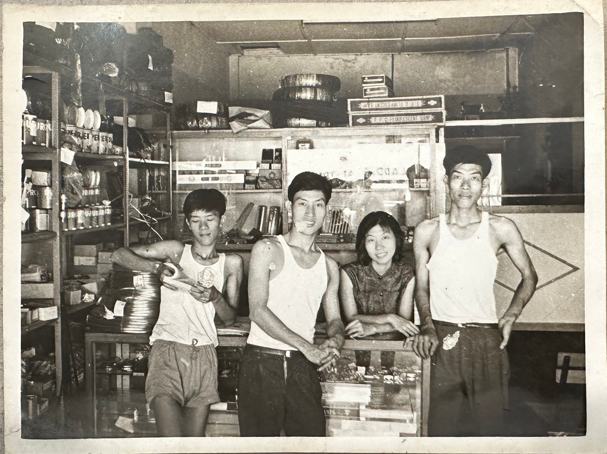 A vintage black and white photograph showing four people standing together inside a  coffee shop with shelves and merchandise in the background.